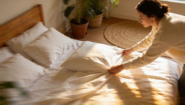 Woman making bed in sunlit minimal bedroom with neutral linen and houseplants
