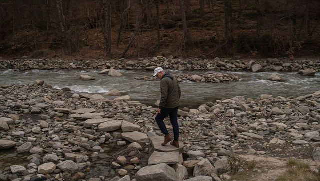 Hiker stepping across rocky river on stepping stones wearing olive jacket and white cap