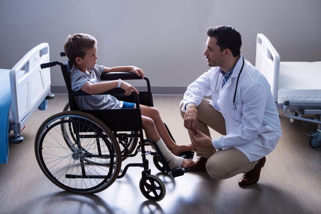 Doctor kneeling and talking to a young child in a wheelchair in a hospital ward. The scene conveys compassion, care, and professional medical attention. Ideal for use in healthcare, pediatric care, medical consultation, and hospital-related content.