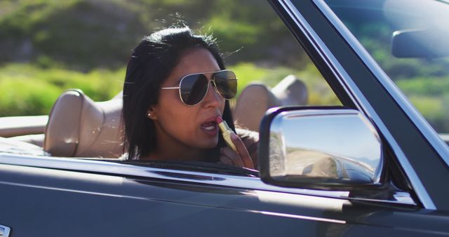 African american woman applying lipstick while looking in the mirror of convertible car. road trip travel and adventure concept