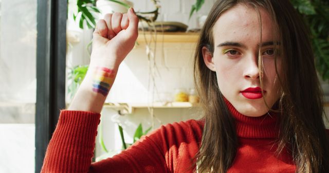Young person confidently raising fist with rainbow painted on wrist, representing LGBTQ+ pride and activism. Wearing red sweater with natural light coming through window, plants in background. Ideal for use in campaigns promoting LGBTQ+ rights, self-expression, diversity, and inclusivity.