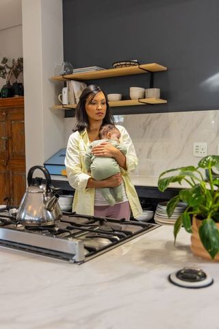 Mother holding sleeping infant while standing at kitchen stove, cradling in calm morning