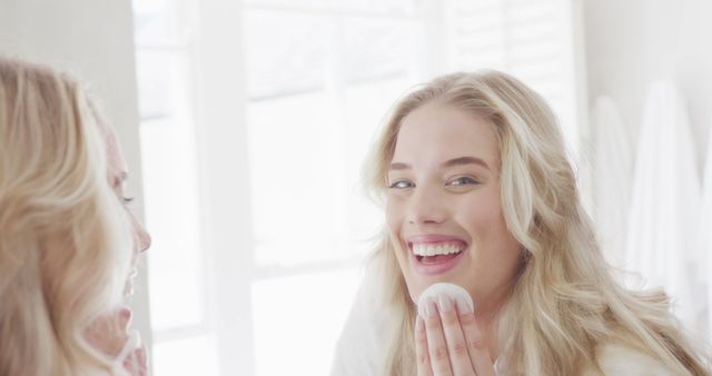 Young woman with blonde hair smiling while applying skincare in a bright bathroom. Perfect for content related to beauty routines, self-care, skincare products, and personal hygiene. The natural light enhances the fresh and healthy look, making it ideal for promoting skincare brands, morning routines, or wellness blogs.