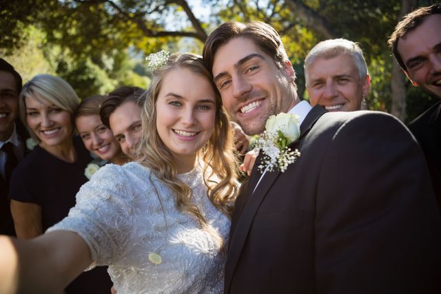 Happy couple posing with guests during wedding in park. Bride and groom smiling with family and friends, celebrating their special day. Ideal for wedding invitations, celebration announcements, and articles on wedding planning.