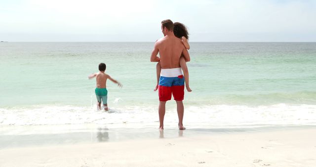 Father stands on shore holding daughter as son runs into shallow waves, displaying family bonding and summer fun. Ideal for articles on parenting, summer vacations, outdoor activities, and beach destinations.