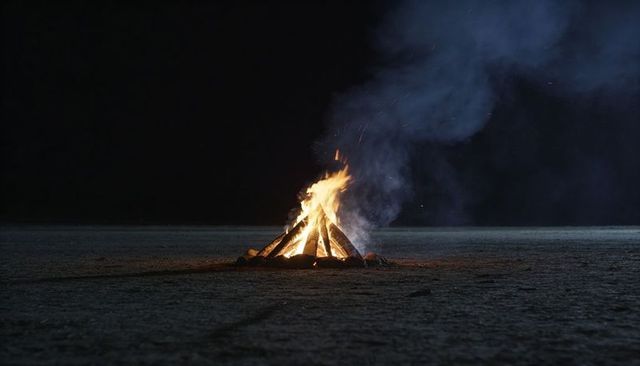 Solitary bonfire burning on salt flat at night with blue smoke and stacked logs