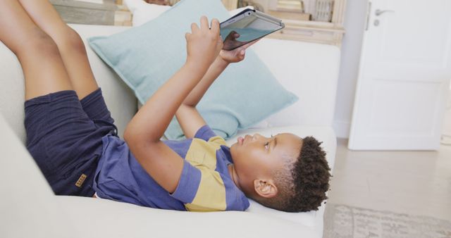 Young boy in blue and yellow casual shirt and shorts lying on a white couch using a tablet. Combines the ideas of a modern lifestyle, leisure time technology use, and comfort at home. Ideal for illustrating concepts like child learning, entertainment, technology in education, and relaxed family living.