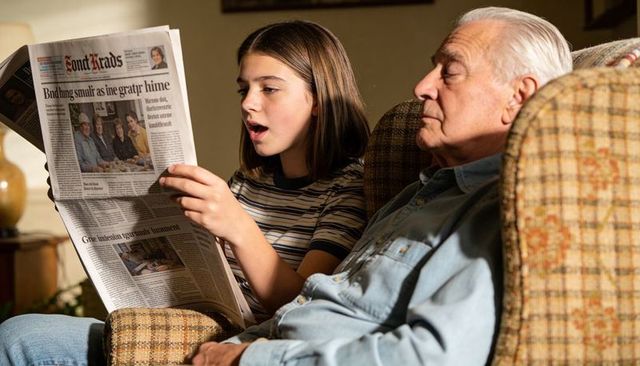 Teen girl reading newspaper aloud to grandfather on cozy living room couch