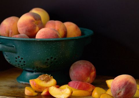 Ripe peaches sitting in teal colander with sliced fruit on wooden tabletop