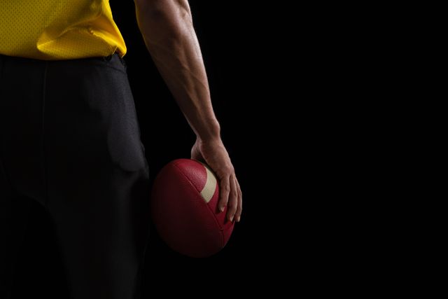 This image shows an American football player holding a ball against a dark background. It is ideal for use in sports-related content, advertisements, promotional materials for football events, fitness and training programs, and articles about teamwork and athletic preparation.