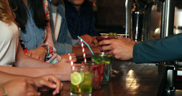 The image shows a small group of friends gathered at a bar counter, enjoying colorful cocktails with straws and garnishes. This photo can be used for lifestyle, nightlife, or socializing themes. Ideal for advertising bars, social venues, or events related to social gatherings.