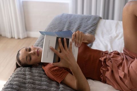 Young woman reading paperback while lying on bed in sunlit minimalist bedroom