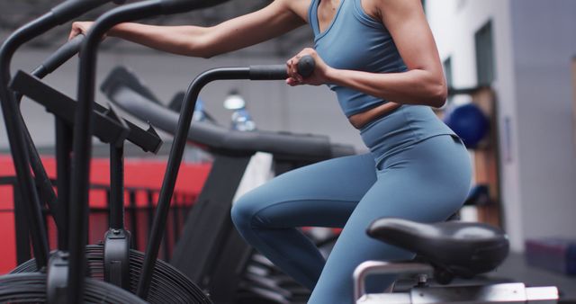 A woman is exercising on a stationary bike machine in a gym, focusing on her workout. She is wearing blue activewear and appears to be in good physical shape. Indoors, the gym is equipped with various workout machines. This image can be used for fitness blogs, workout tips, gym advertisements, health and wellness promotions, or athletic apparel marketing.