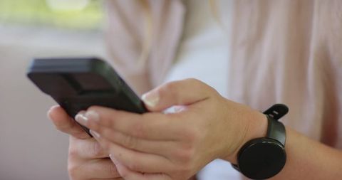 Woman holding smartphone with case, checking black smartwatch by window in living room