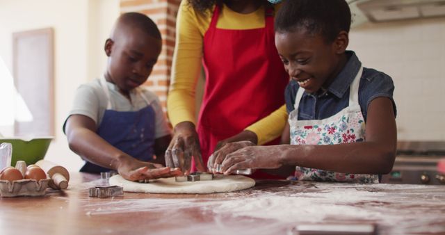 Mother supervising her two children carving cookie shapes out of dough in a kitchen setting. Children are wearing aprons, flour spread on the table. Ideal for use in family-oriented marketing, cooking classes, educational materials, advertisements showcasing family moments, or content on parenting and child development.