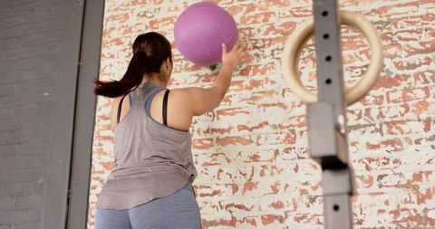 Woman throwing purple medicine ball at whitewashed brick wall during functional training