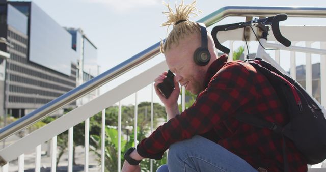 Young man with headphones crouching outdoors, using smartphone. Urban background with buildings and greenery. Ideal for themes around technology, communication, youth lifestyle, and outdoor activities.