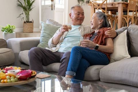 Senior asian couple relaxing on sofa sharing drinks and fresh fruit in cozy living room