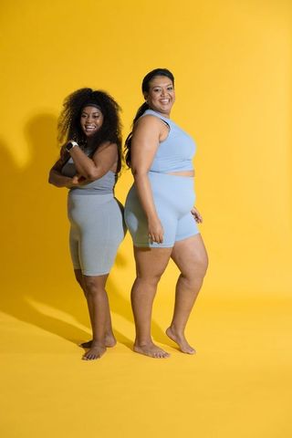 Confident diverse women posing back-to-back in pastel activewear on yellow background