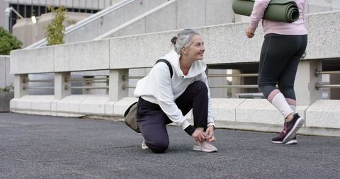 Mature women tying sneakers in urban plaza before yoga, active senior wellness lifestyle