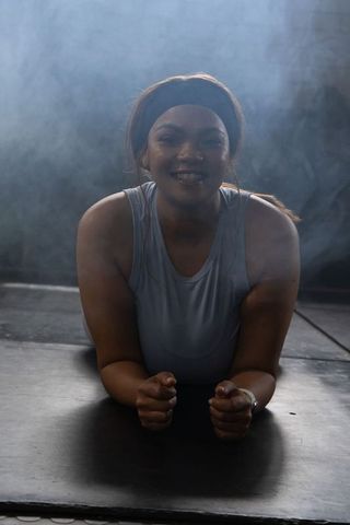 African american woman holding forearm plank on dark mat, smiling in smoky studio workout