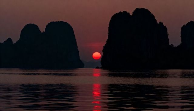 Crimson sun sinking between karst cliffs with mirror red reflection on calm sea at dusk