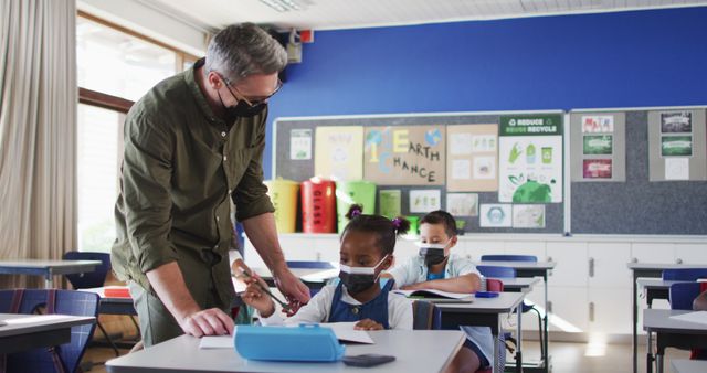 Diverse male teacher helping schoolgirl sitting in classroom, all wearing face masks. children in primary school during coronavirus covid 19 pandemic.