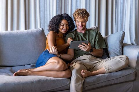 Mixed-race couple relaxing on gray sofa using tablet together in cozy modern living room