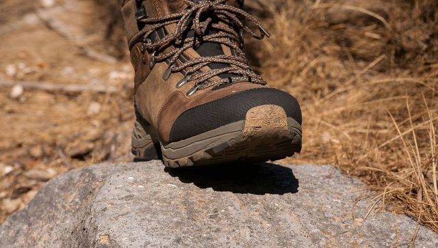 Brown leather hiking boot balancing on rock with rubber toe cap and mud-streaked lugged sole