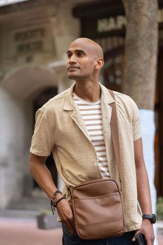 Man standing on city sidewalk wearing beige shirt with brown leather crossbody bag checking phone