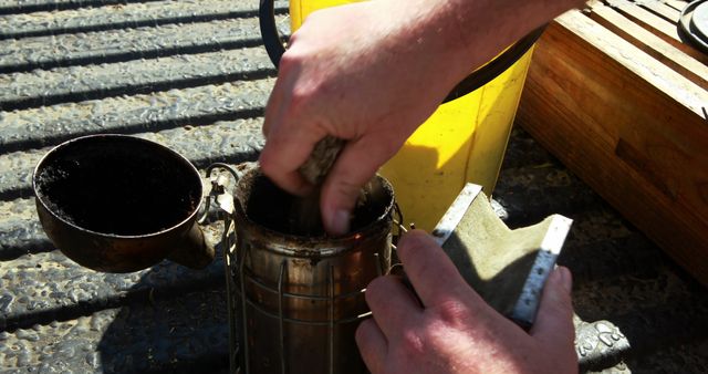 Close-up of a beekeeper's hands preparing beekeeping equipment, including a bee smoker and other tools, for inspecting a beehive. Ideal for articles, tutorials, and educational content about beekeeping practices, apiary management, and agricultural lifestyle.