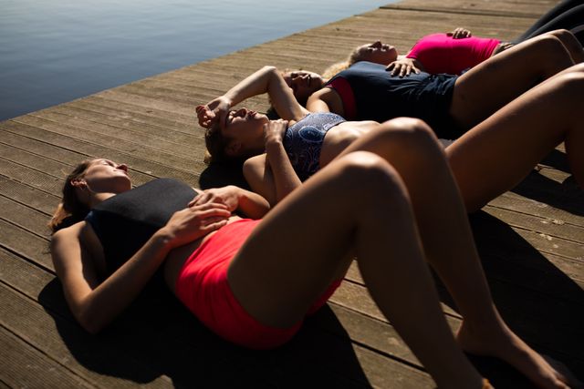 High angle side view of a rowing team of four Caucasian women wearing sportswear and training on a jetty by the river, lying down taking a break and relaxing with river in the background.