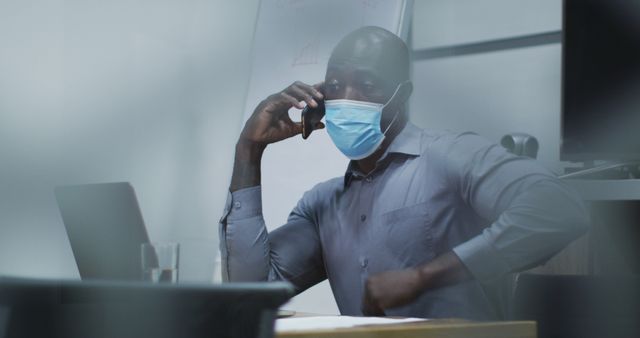 African american businessman in face mask talking on smartphone, using laptop in conference room. business professional working in modern office during covid 19 coronavirus pandemic.