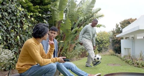 Young friends relaxing on backyard bench while friend juggling soccer ball on lawn
