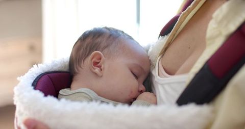 Mother cradling sleeping baby in soft carrier by window