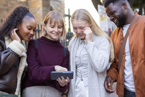 Diverse friends laughing and pointing at smartphone while walking city street
