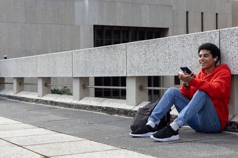 African american teen sitting on urban plaza wall wearing red hoodie holding smartphone