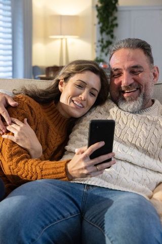 Couple cuddling on couch wearing cable knit sweaters, laughing while looking at smartphone