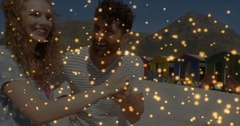 Laughing couple embracing on sunlit beach with colorful huts and golden bokeh sparkles