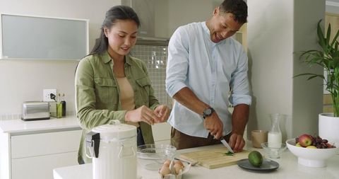 Diverse couple cooking together on kitchen island, cracking eggs and chopping herbs