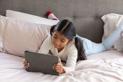 School-age girl lying on bed using tablet for remote learning and relaxed screen time