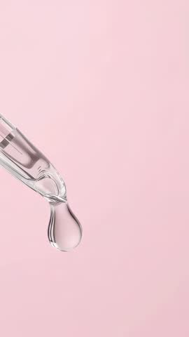 Vertical close-up of serum drop forming and falling from glass dropper on pink background