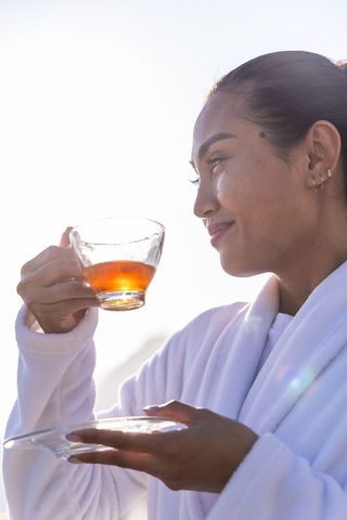 Asian woman enjoying morning tea in white bathrobe holding glass teacup on balcony