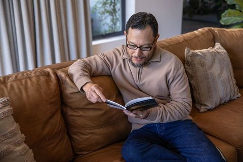 Mature asian man relaxing on leather sofa reading paperback book and pointing at page