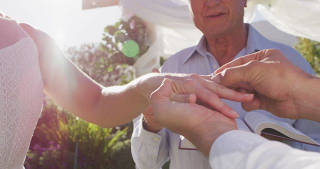 Intimate moment as bride and groom exchange rings during sunlit outdoor wedding ceremony. Ideal for event planning advertisements, bridal magazines, wedding invitations, blog posts about wedding trends, or social media promotions for wedding services.