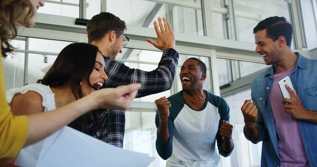This image showcases a diverse group of young professionals celebrating in an office. They appear excited and energetic, emphasizing the importance of teamwork and success in a collaborative business setting. Ideal for advertising company culture, teamwork, success stories, or motivational content.
