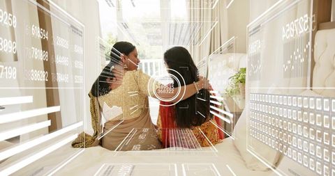 Indian mother and daughter embracing on bed in sarees with digital hud overlay, sunlight