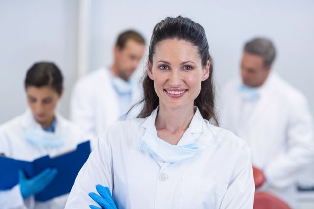 Dentist standing confidently with arms crossed in a dental clinic, smiling at the camera. Background shows other dental professionals engaged in their tasks. Ideal for use in healthcare, dental care, and medical team-related content. Can be used for promoting dental services, healthcare teamwork, and professional medical environments.