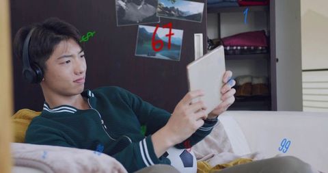 Teenager relaxing on couch wearing headphones and using tablet with soccer ball nearby