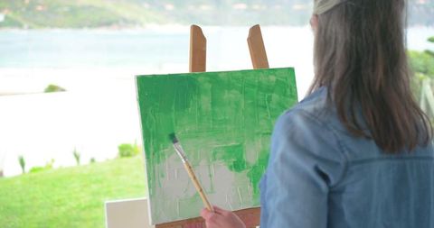 Woman painting green abstract canvas by lake holding brush at wooden easel outdoors